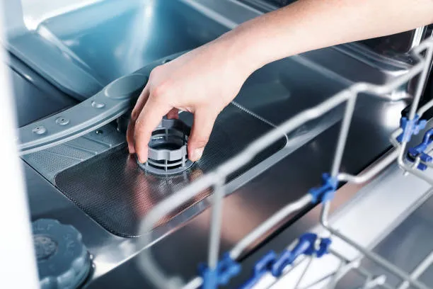 A hand removing the circular grey drain filter from the bottom of a dishwasher for cleaning.