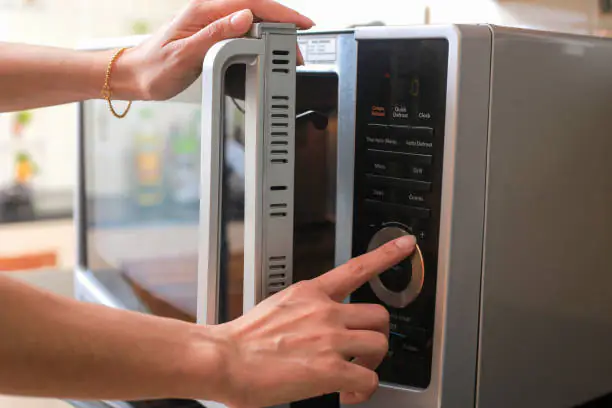 A close-up of a person's hand pressing buttons on a modern silver microwave control panel to start a cooking cycle in a kitchen.